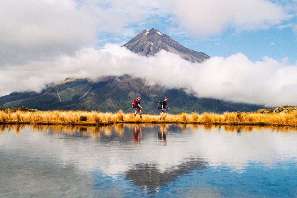 两个人在湖边散步，湖后面有一座被白云环绕的火山